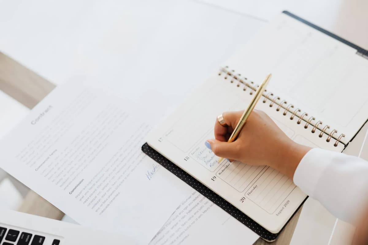 A hand writing in a planner beside a printed contract on a desk
