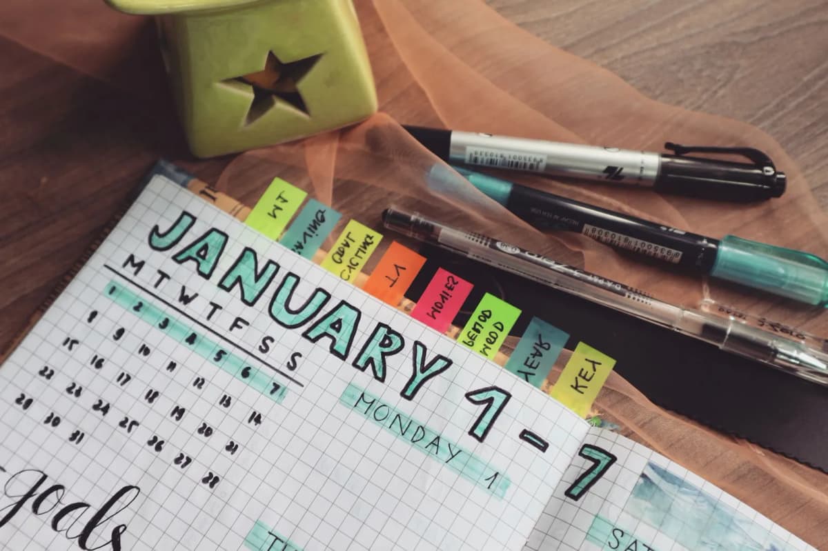 A planner with written goals and colored tabs open on a wooden desk beside pens