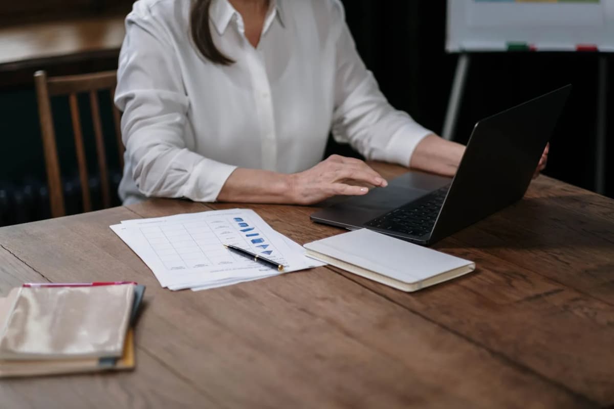 A person at a wooden table checking printed account figures against a laptop