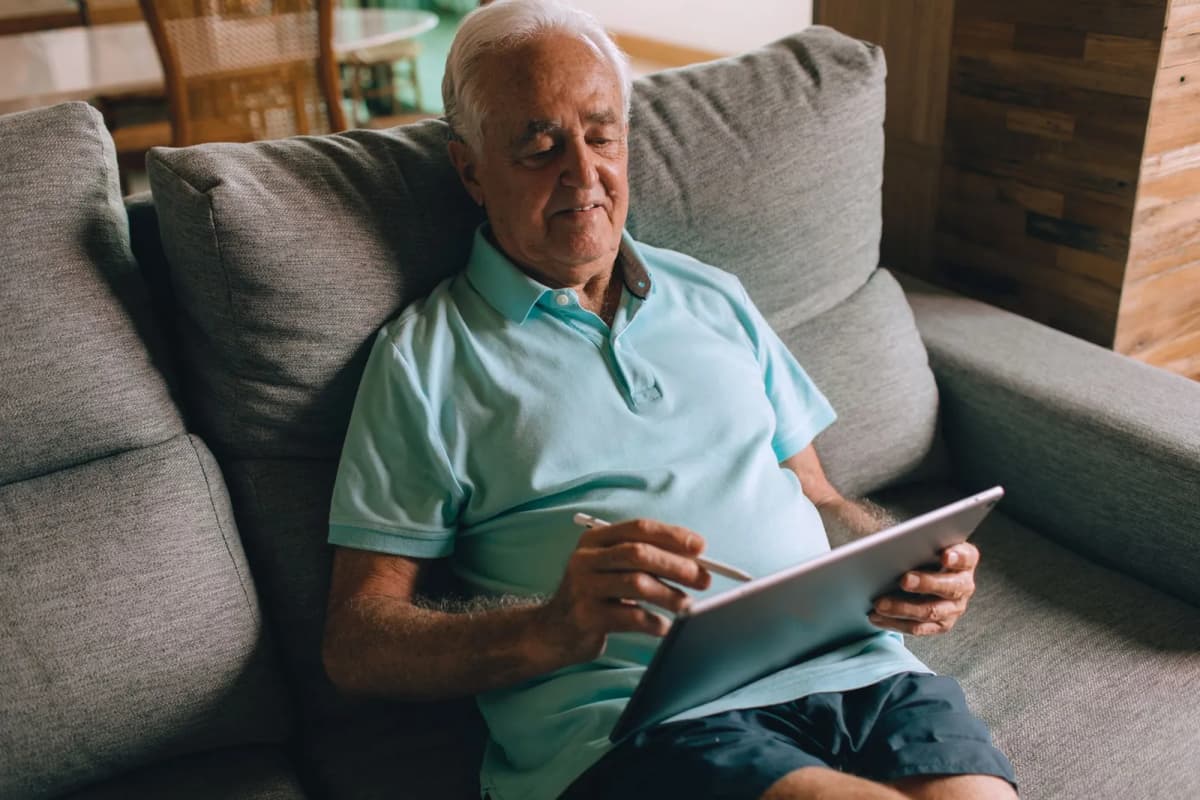 An older man sitting on a couch at home, using a tablet with a stylus