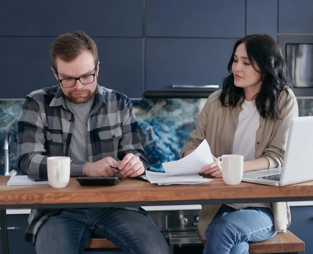 A couple at their kitchen table going through bills with a calculator and a laptop
